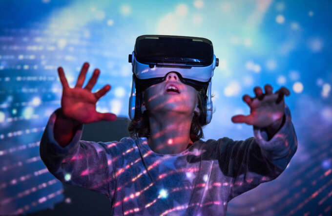 Amazed little child looking up with opened mouth and raising hands while exploring cyberspace in VR goggles standing near wall with colorful projection